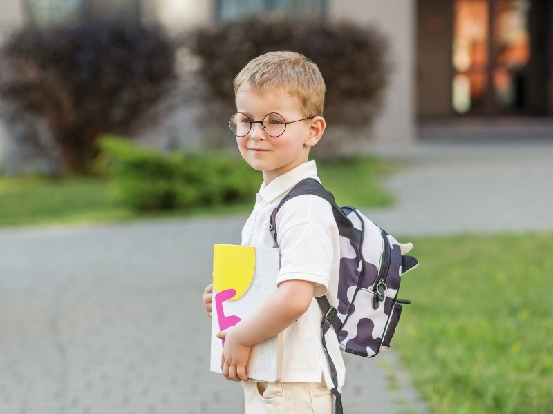 Young boy with glasses and bookbag in front of school, highlighting the back to school checklist for parents.