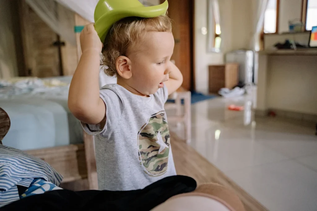 A young boy playing indoors, putting on a green plastic hat in a cozy bedroom setting. Image complements the blog, how to potty train for daycare