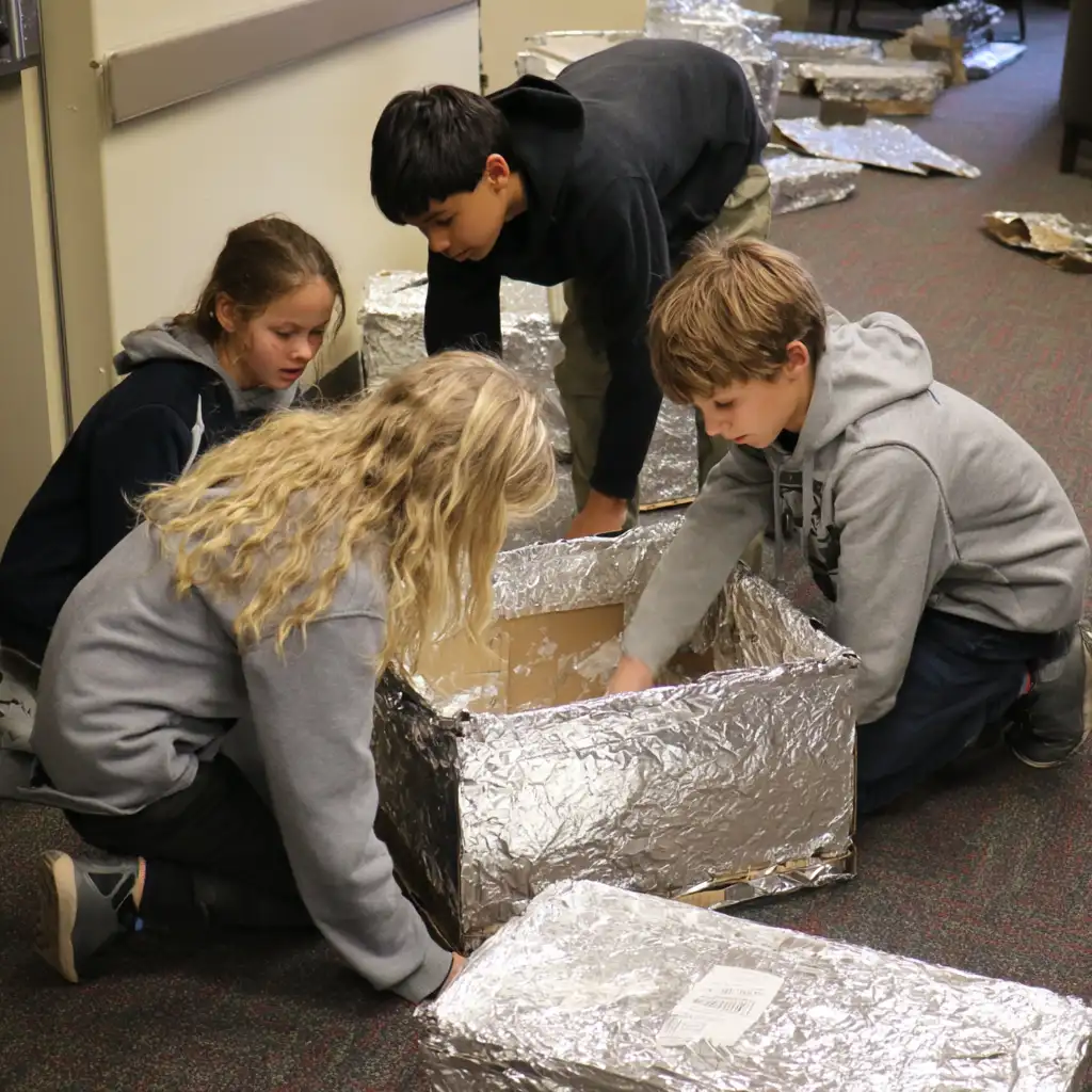 Children crafting an oven out of a pizza box, aluminum foil and plastic wrap. 